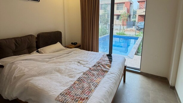 A bedroom with a bed, a ceiling fan, an air conditioning unit, a window with curtains, and a view of buildings and a swimming pool.