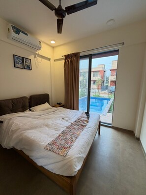 Una camera da letto con un letto, un ventilatore a soffitto, un condizionatore d'aria, una finestra con tende e vista su edifici e una piscina.