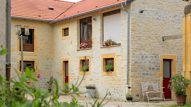 A stone building with multiple windows, some with shutters, and a red door. There are potted plants and a white bench outside.