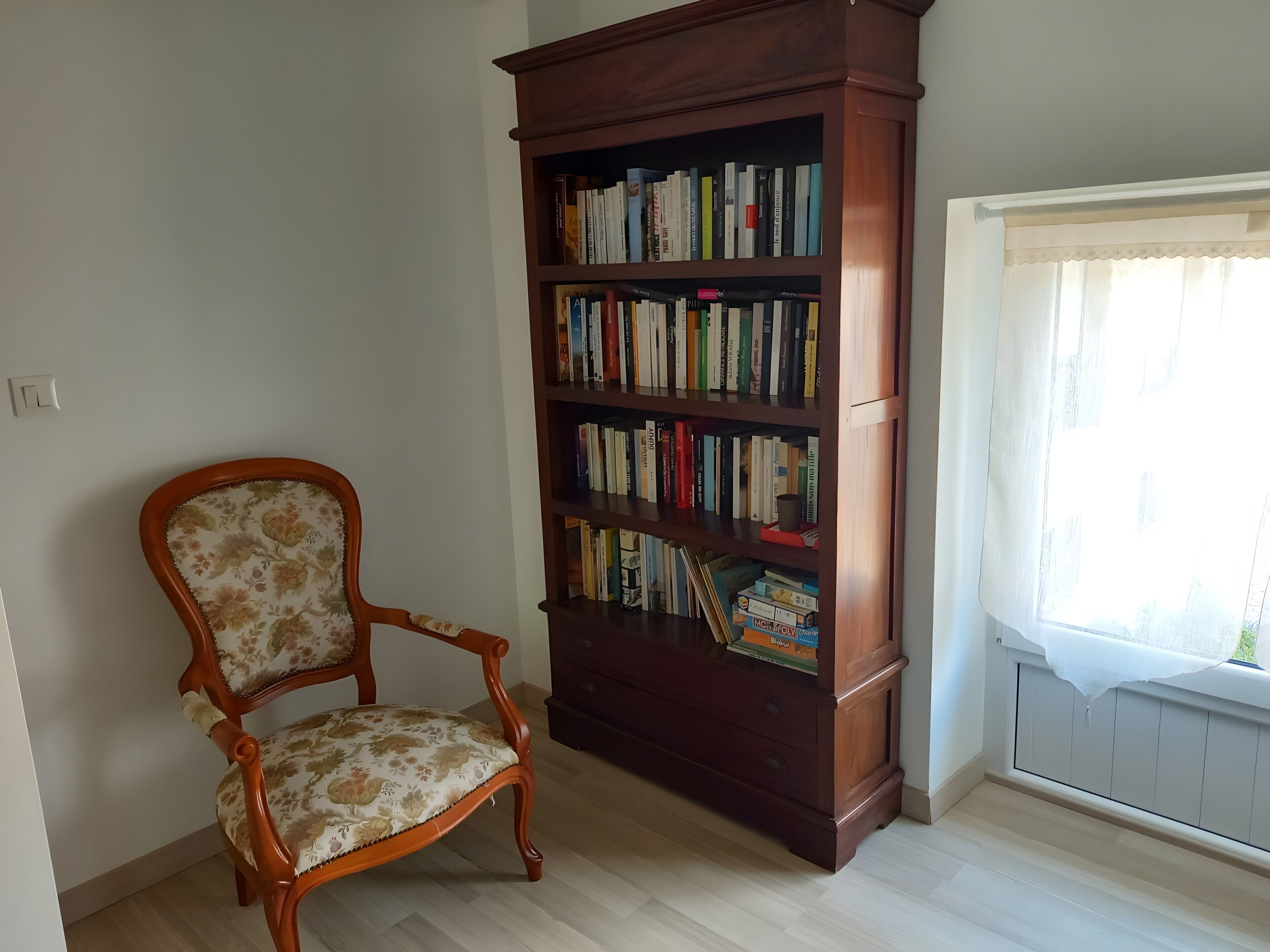 Een kamer met een houten boekenkast vol boeken, een fauteuil met bloemenmotief en een raam met een witte gordijn.