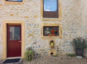 Ein Steinhaus mit roter Tür, einem Fenster mit Blumenkasten und einem kleinen Fenster mit gelbem Eimer.