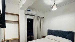 A bedroom with a bed, a TV mounted on the wall, a wooden bench, and a ceiling light. - LBC Hotel (Sarandë)