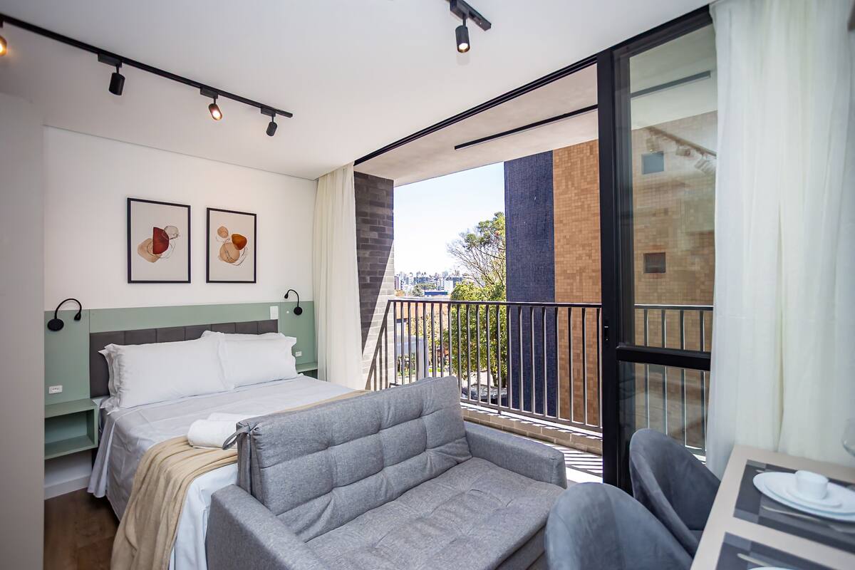 A modern hotel room with a bed, grey sofa, and a balcony with a city view.