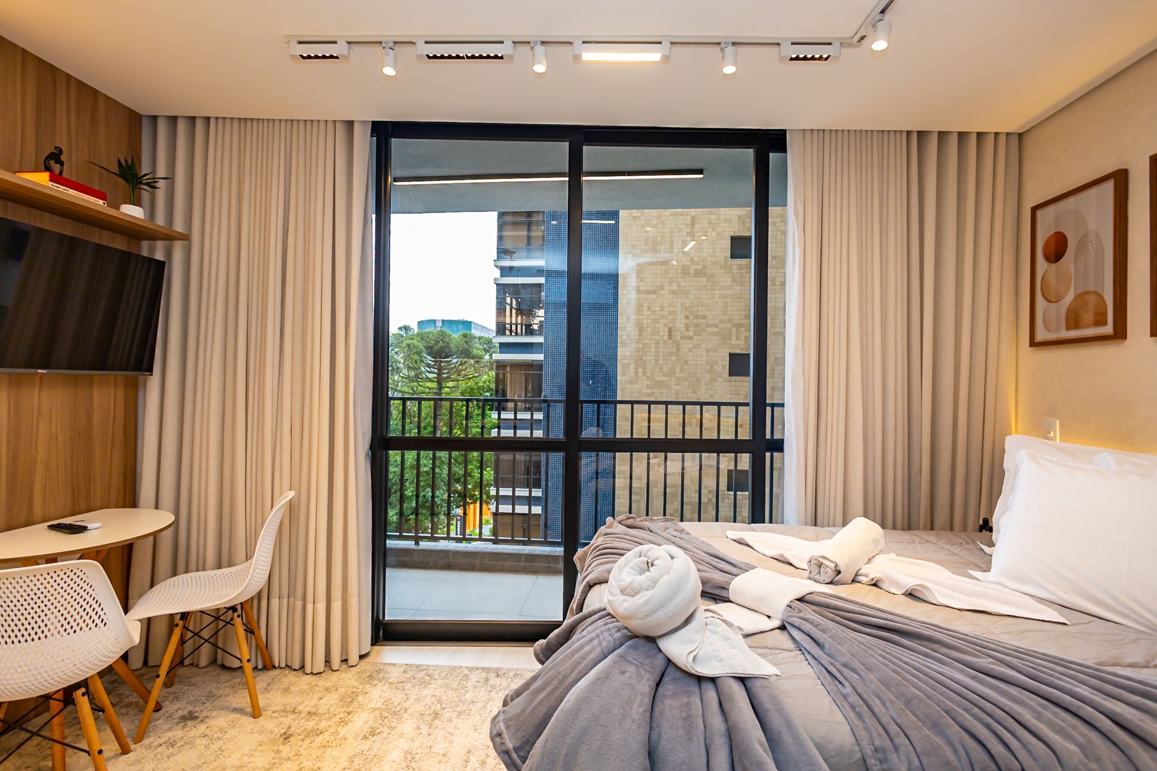 Habitación de hotel con cama, escritorio, silla, televisión y balcón con vistas a edificios.