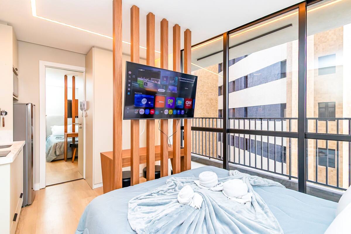 A modern hotel room with a large bed, a TV mounted on a wooden panel, a balcony with a view of the building, and a small kitchen area.