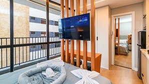A modern hotel room with a large TV, a balcony with a view of the city, a bed with a grey headboard, and a wooden partition. - Oxygen Batel (Curitiba)
