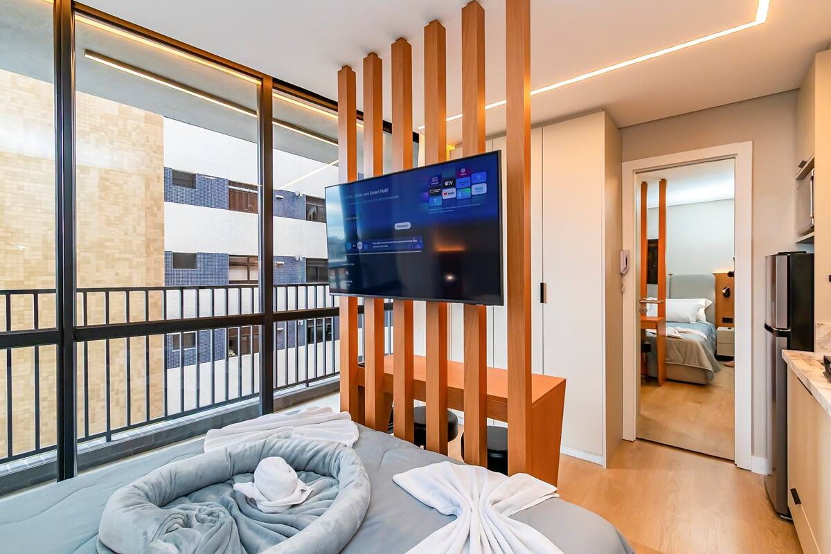 A modern hotel room with a large TV, a balcony with a view of the city, a bed with a grey headboard, and a wooden partition.
