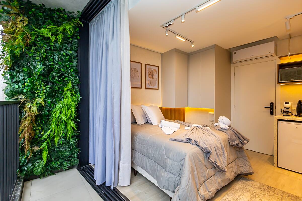 A modern hotel room with a bed, a small kitchenette, a wall-mounted air conditioner, and a curtain.