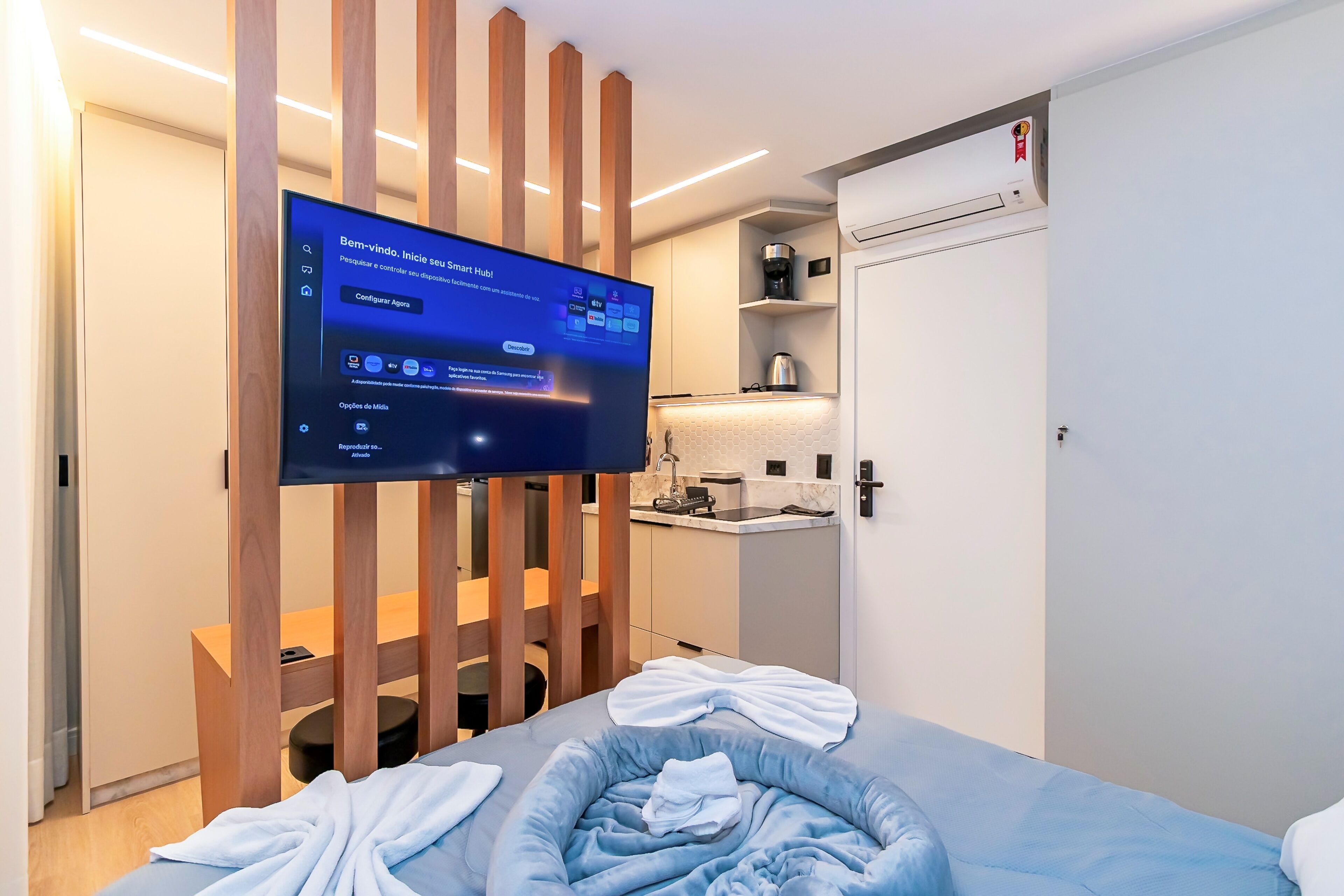 A modern hotel room with a bed, a TV mounted on a wooden partition, a small kitchenette, and a door with an air conditioning unit.