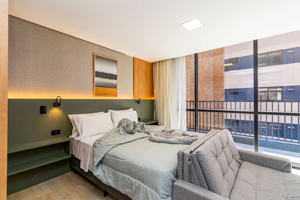 A modern hotel room with a bed, a sofa, a large window, and a painting on the wall.