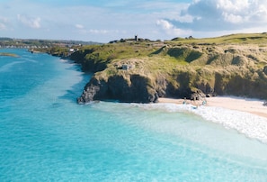 A coastal landscape with clear turquoise waters, grassy hills, and a distant building on a hill. - Acton Castle, Marazion, Cornwall (Praa Sands)