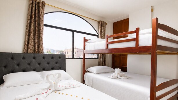 Una habitación de hotel con una cama grande, dos camas más pequeñas, una ventana con cortinas, una lámpara de techo y vistas a edificios.