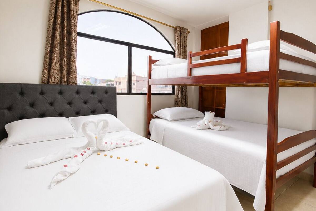 Una habitación de hotel con una cama grande, dos camas más pequeñas, una ventana con cortinas, una lámpara de techo y vistas a edificios.