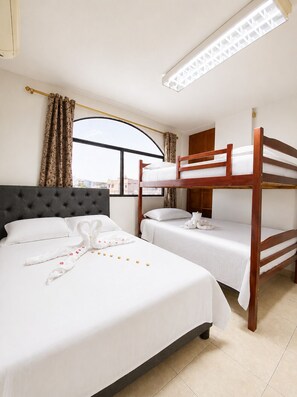 A hotel room with a large bed, two smaller beds, a window with curtains, a ceiling light, and a view of buildings outside. - Galapagos Home (Puerto Baquerizo Moreno)