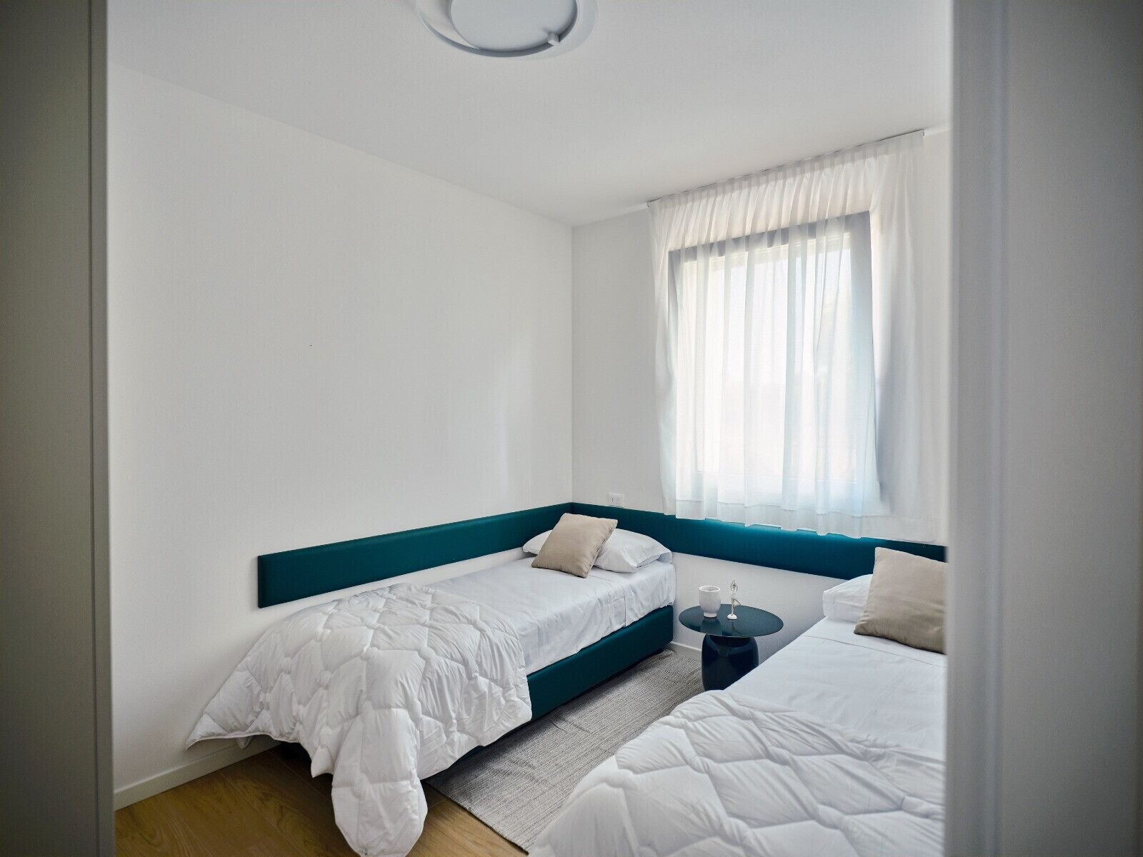 Un dormitorio con dos camas individuales, una mesita y una ventana con cortinas de gasa.