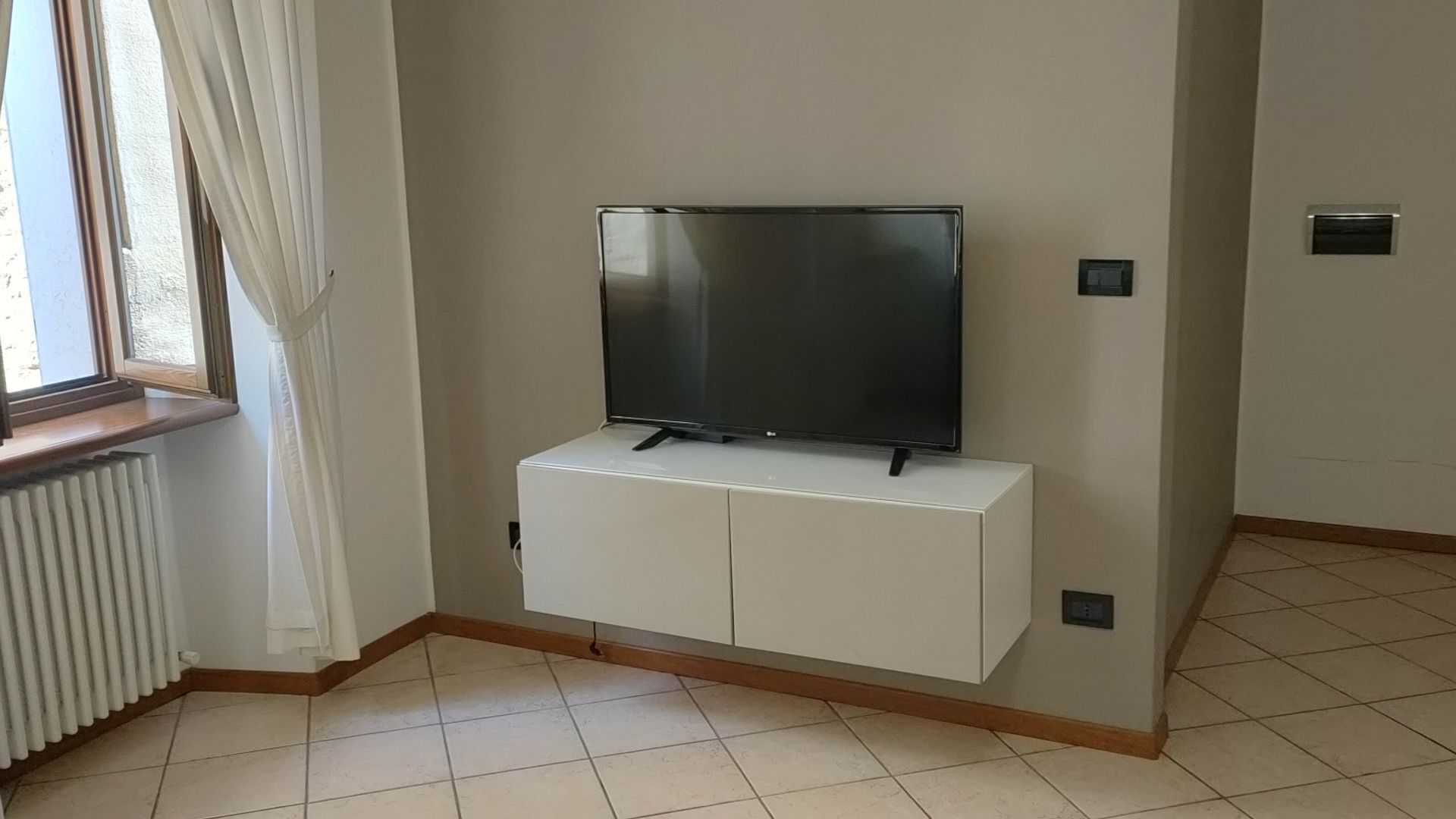 A room with a TV mounted on a white cabinet, a window with curtains, and a radiator.