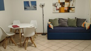 A living room with a blue sofa, white chairs, a small round table with a red book, and two artworks on the wall. - Appartamento con 1 Camera da letto (Riva del Garda)