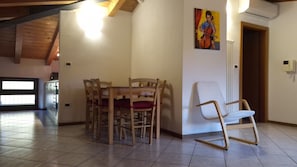 A dining area with a wooden table and chairs, a white chair, a painting on the wall, and a kitchen in the background. - Appartamento Grande con 1 Camera da Letto (Riva del Garda)