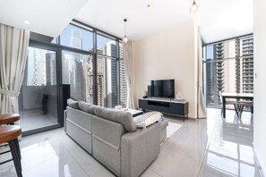 Ein modernes Wohnzimmer mit grauem Sofa, einem Flachbildfernseher und einem Essbereich mit Tisch und Stühlen. Große Fenster bieten Blick auf hohe Gebäude.