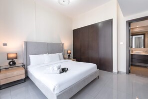 Ein modernes Hotelzimmer mit einem großen Bett, Nachttischen inklusive Lampen, einem dunklen Kleiderschrank und einem durchgegangene Tür erkennbaren Badezimmer.