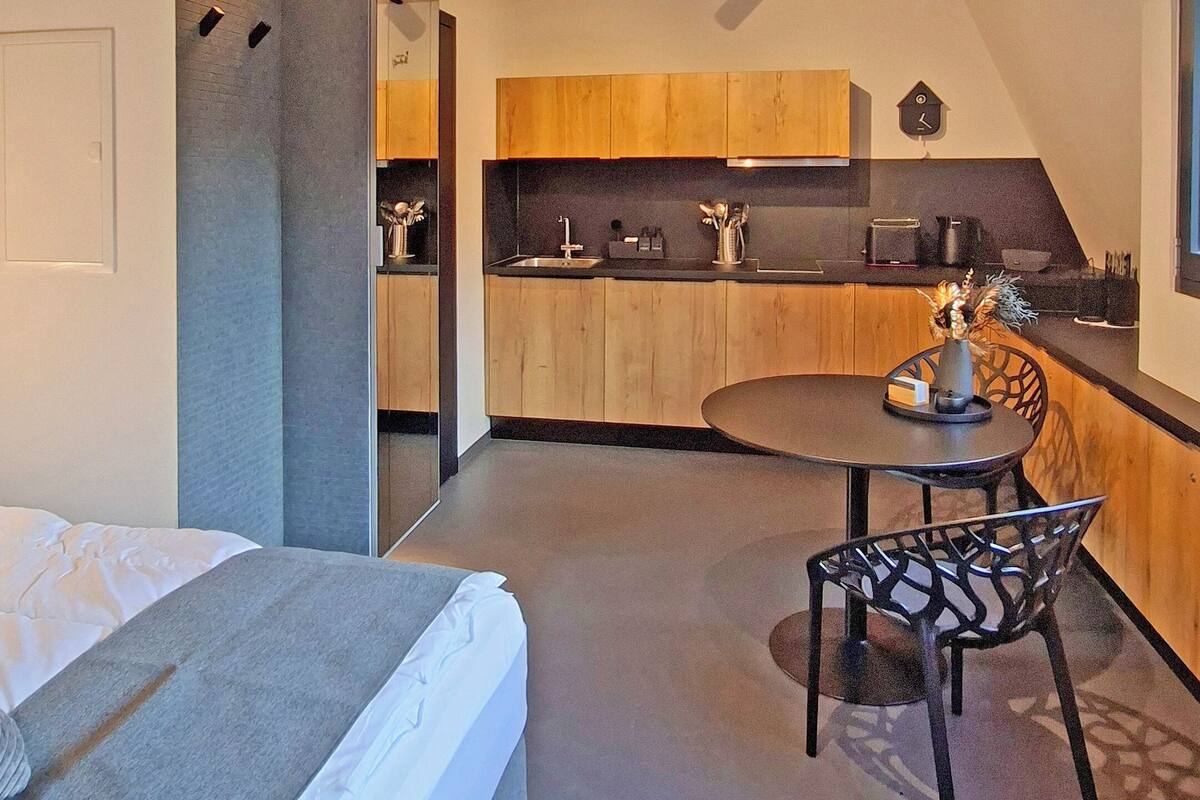 A modern bedroom with a bed, a small table, and a chair. There is a kitchenette with wooden cabinets and a sink.