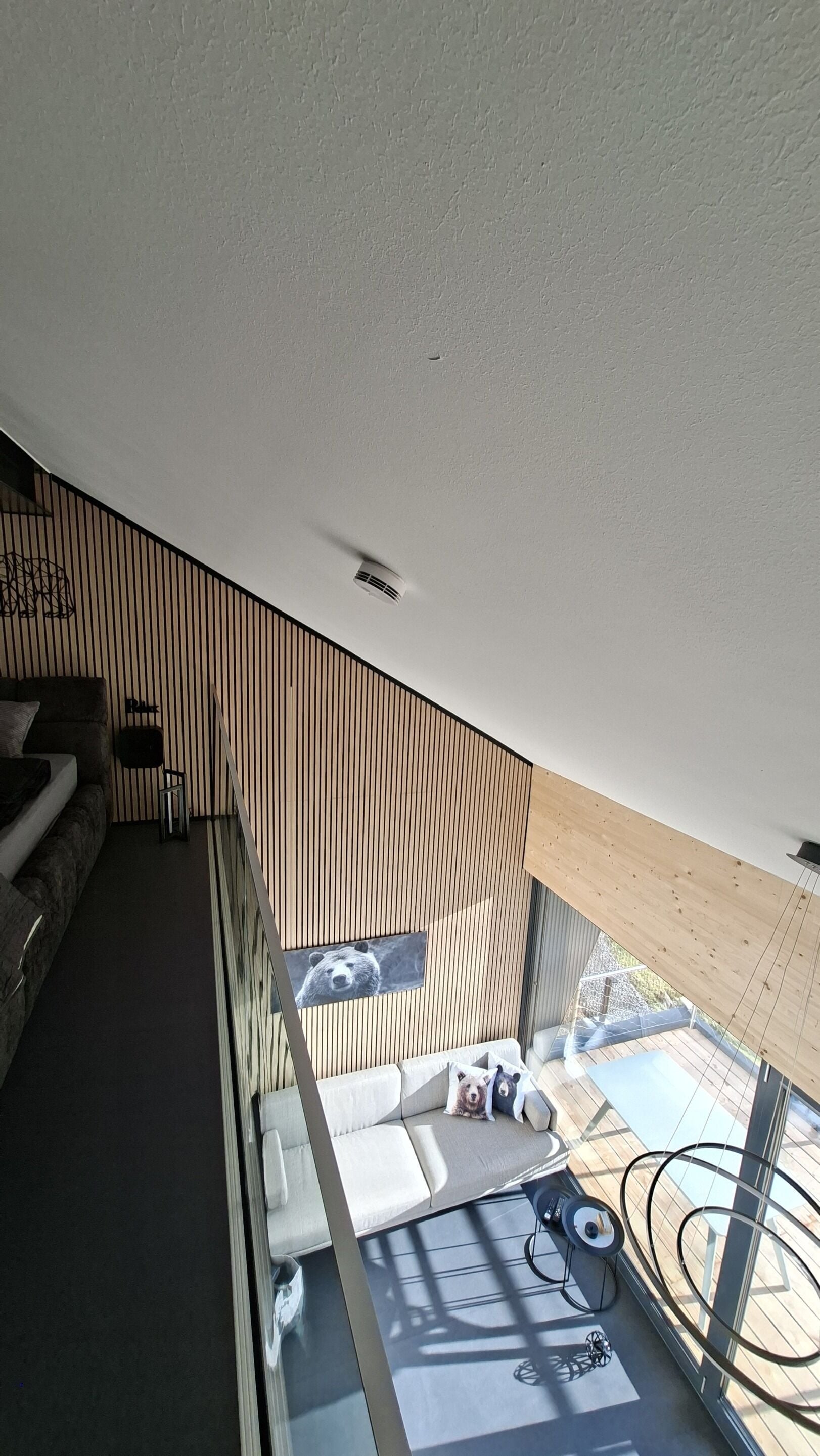 Een modern interieur met een schuine plafond, een houten wandpaneel, een bank en een glazen balustrade met uitzicht op een terras.