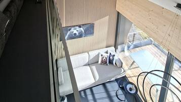 Een modern interieur met een schuine plafond, een houten wandpaneel, een bank en een glazen balustrade met uitzicht op een terras.