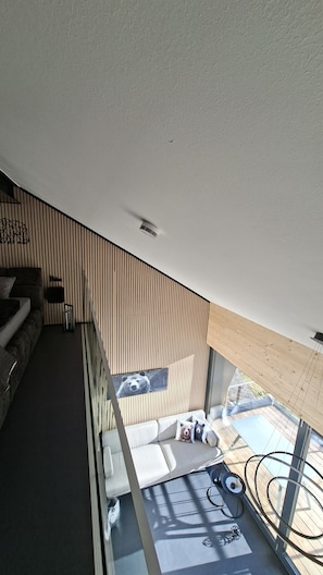 Un interior moderno con techo inclinado, panel de madera, sofá y barandilla de vidrio con vistas a una terraza.