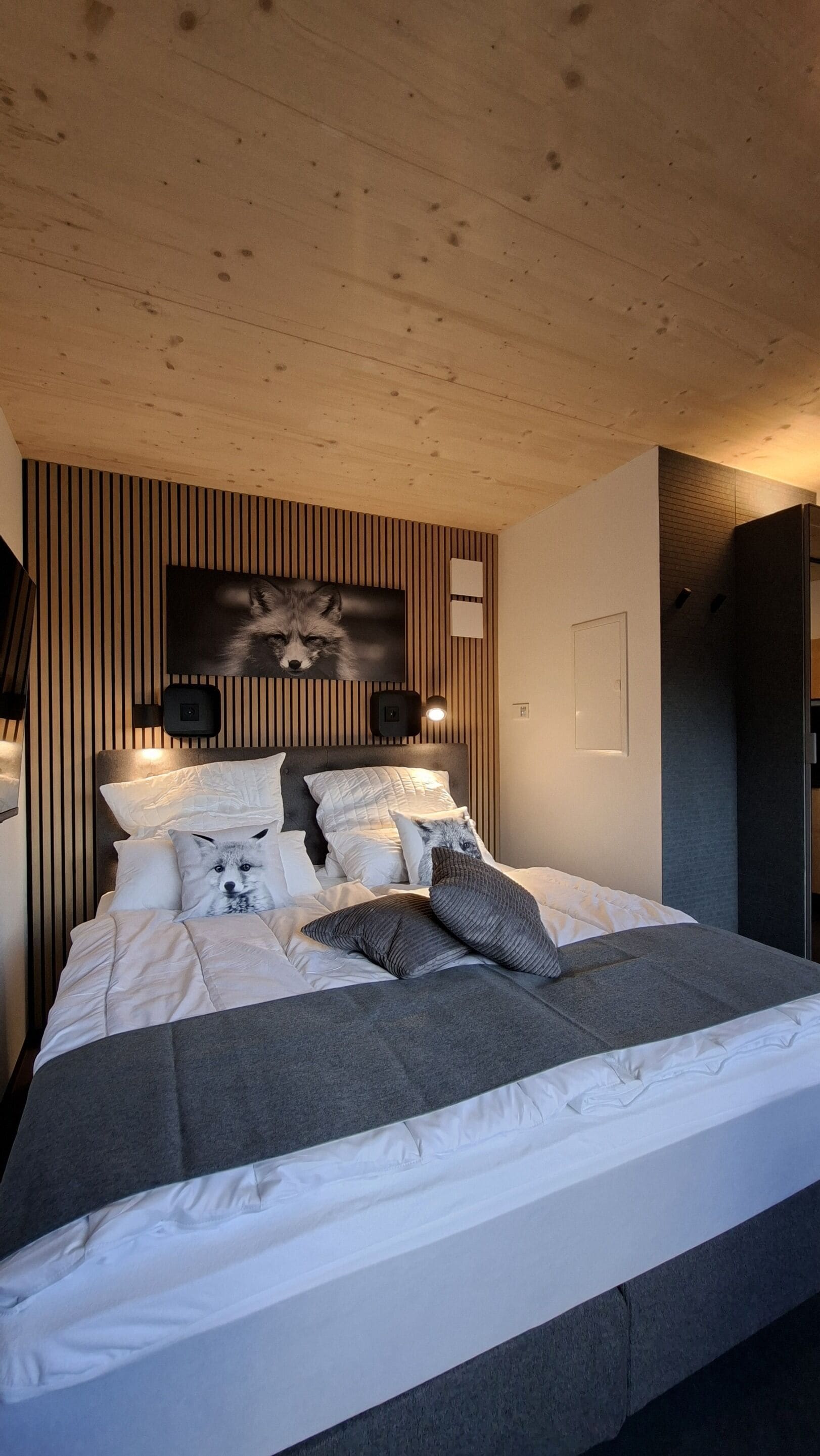 A modern bedroom with a wooden ceiling, a bed with white and gray bedding, a wall-mounted TV, and a wooden panel wall.