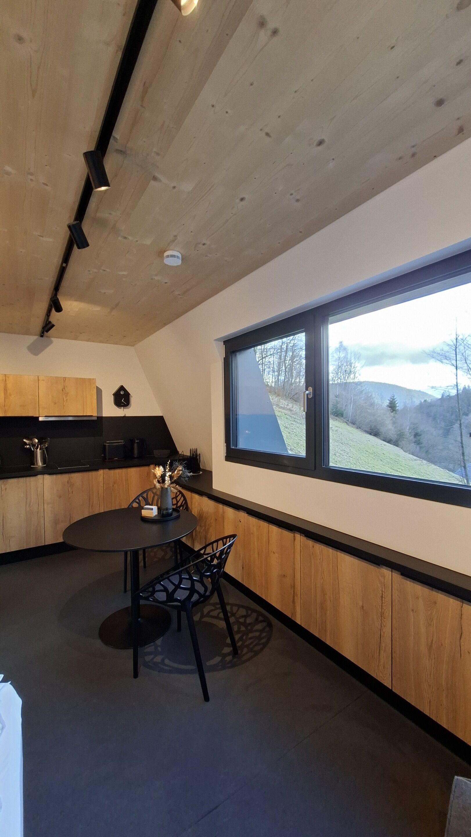 A modern kitchen with wooden cabinets, a round dining table, and a window with a view of a hillside.