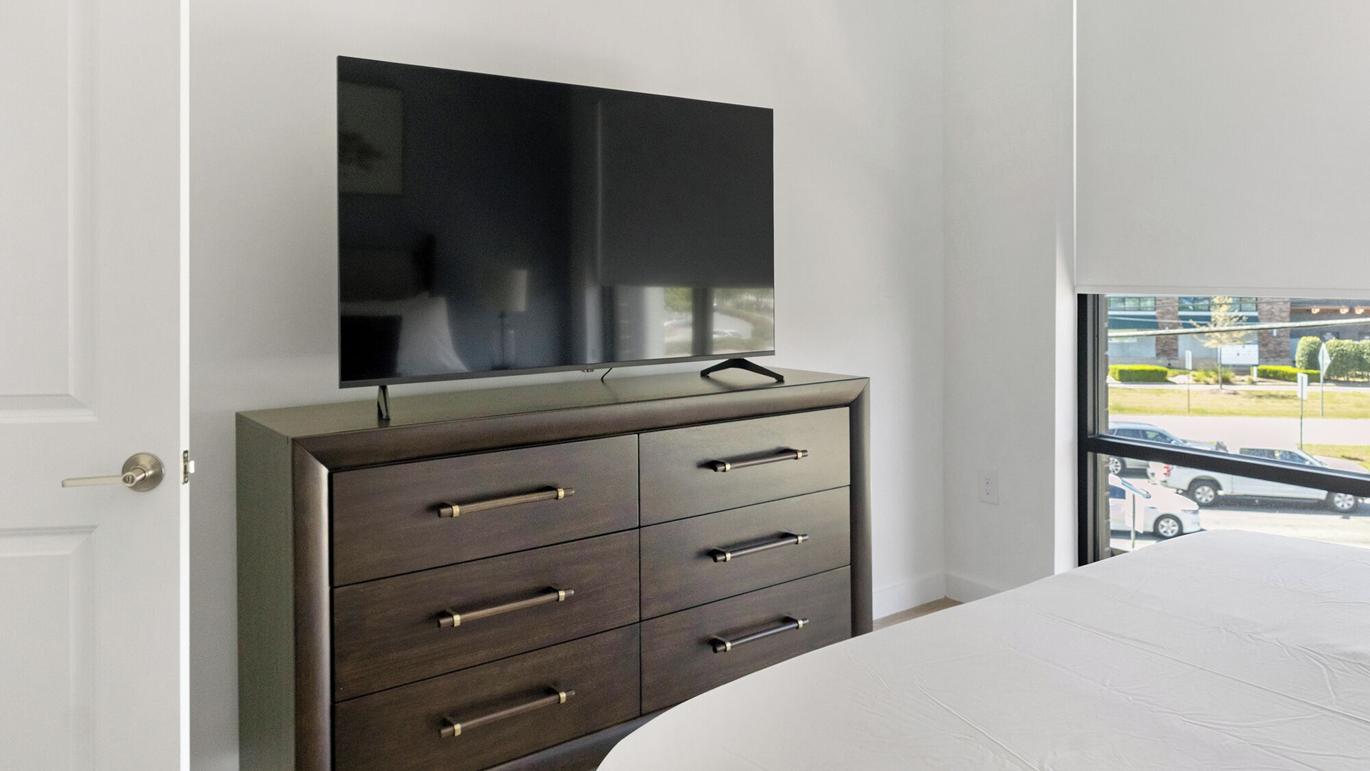 A bedroom with a bed, a dresser, a TV mounted on the wall, and a window with a view of the outside.