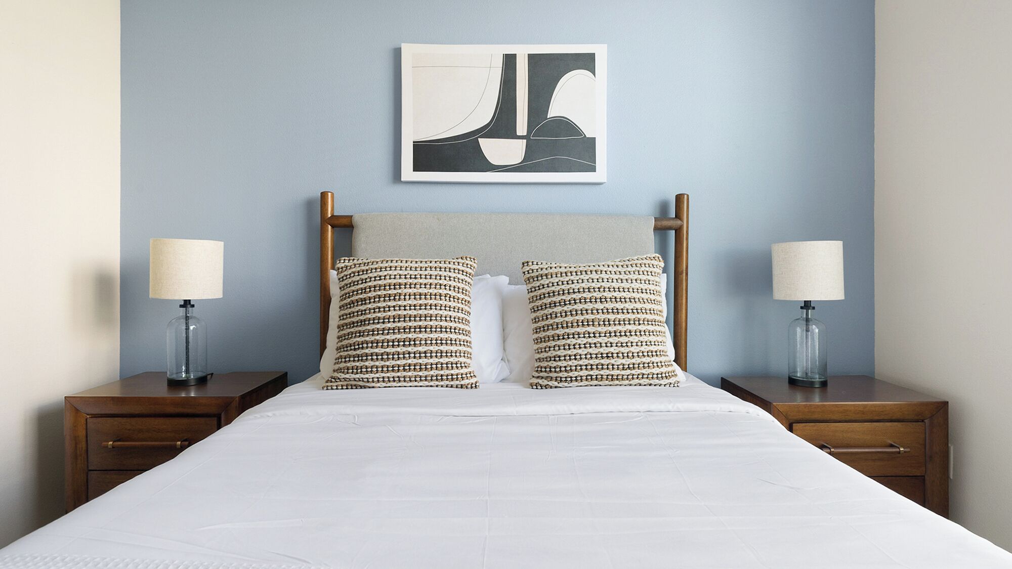 Una cama bien hecha con edredón blanco, dos almohadas estampadas y un cabecero de madera. Hay dos mesitas de noche con lámparas y una obra de arte enmarcada sobre la cama.