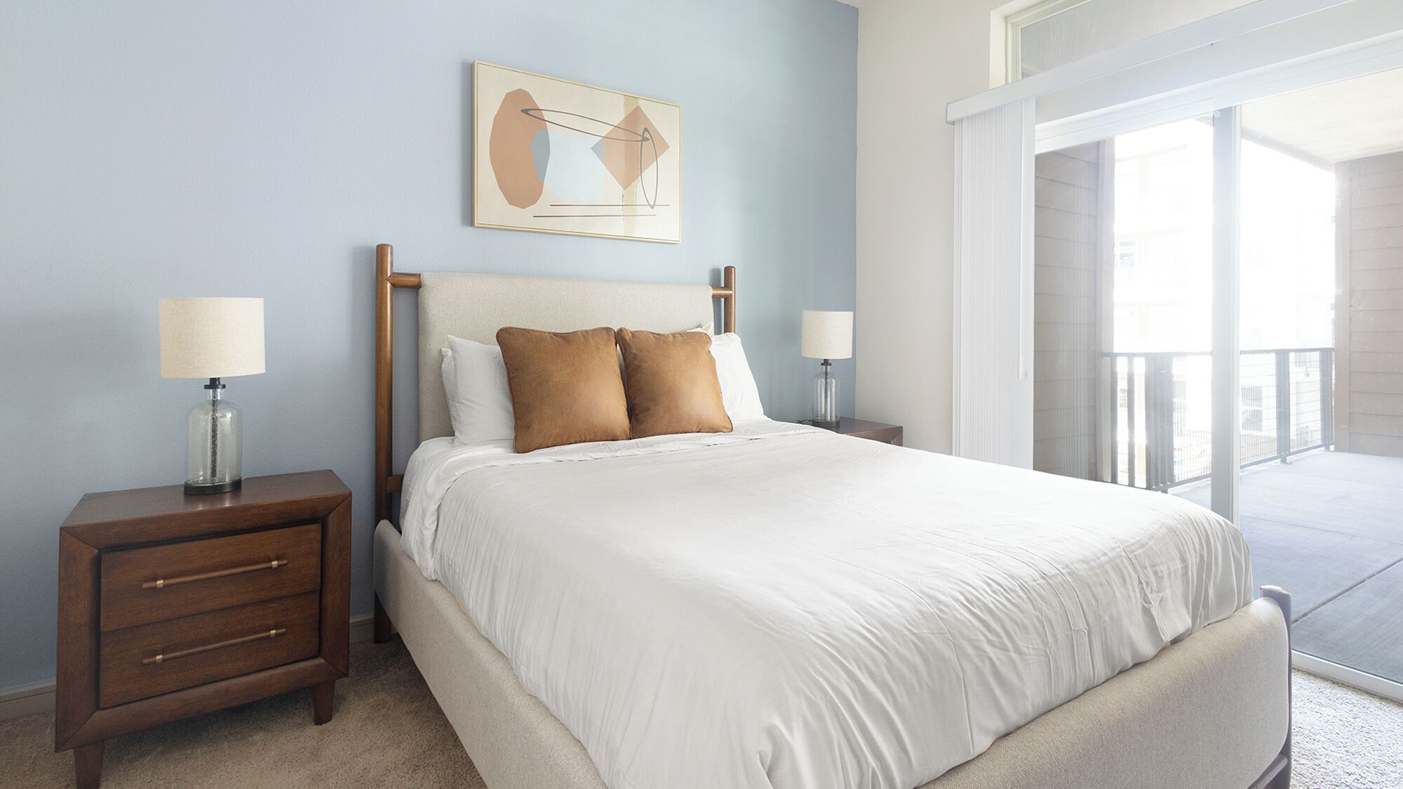 A bedroom with a bed, two bedside tables, a nightstand, a framed picture on the wall, and a balcony with a railing.