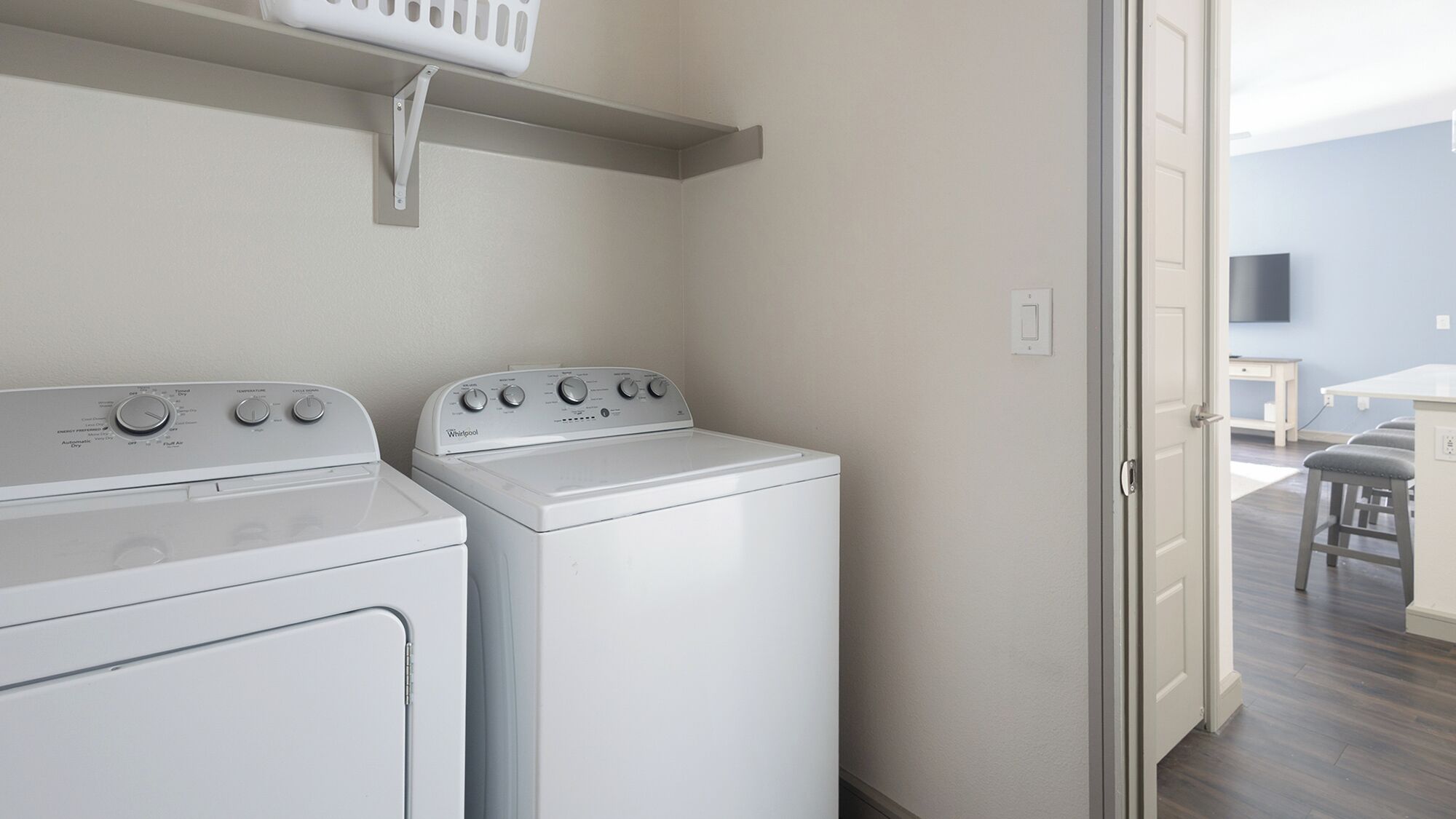 A laundry room with two white washers and dryers, a small shelf above the dryers, and a door leading to a living area with a TV and a table.