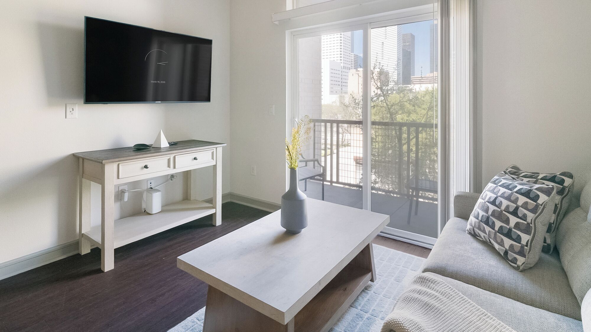 Ein modernes Wohnzimmer mit Flachbildfernseher, weißem Konsolentisch, Couchtisch, einer Couch mit gemusterten Kissen und einem Balkon mit Blick auf Gebäude.