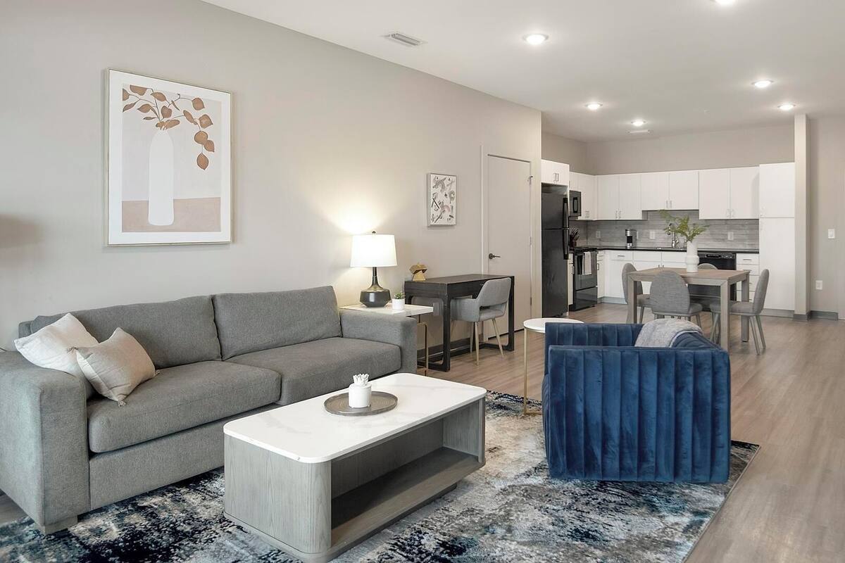 Un salon moderne avec un canapé gris, un fauteuil bleu et une table basse. On aperçoit au fond une cuisine avec des meubles blancs et un coin repas.