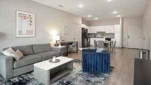 A modern living room with a grey sofa, a blue ottoman, and a coffee table. There is a kitchen in the background with white cabinets and a dining area. - Landing Furnished Apartments Harborside - Memphis (Memphis)