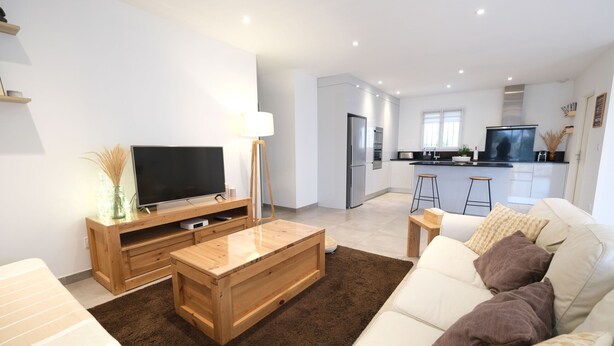 A modern living room with a flat-screen TV, wooden coffee table, and a sofa with patterned cushions. A kitchen with a refrigerator and stove is visible in the background. - Holiday rental with pool in the Alpilles for 6 people (Plan-d´Orgon)