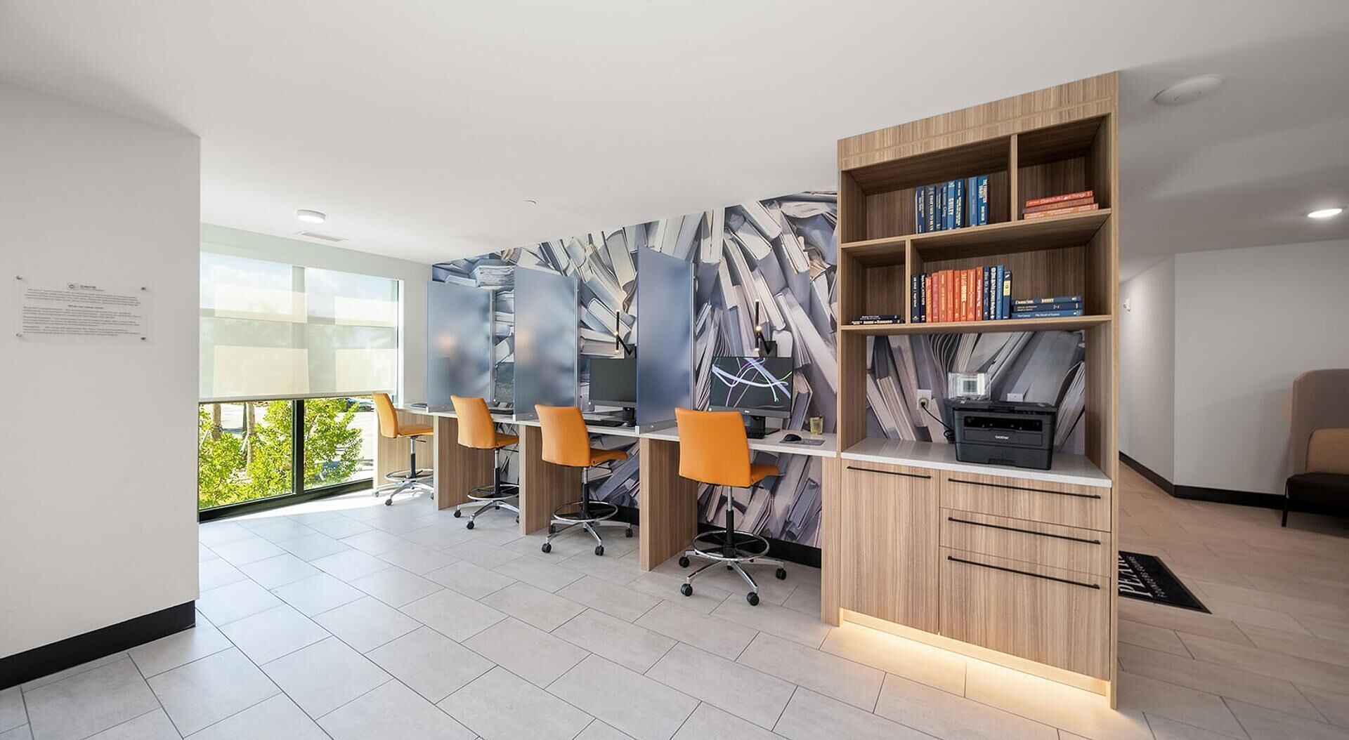 A modern office space with desks, chairs, a bookshelf, and a view of greenery outside.