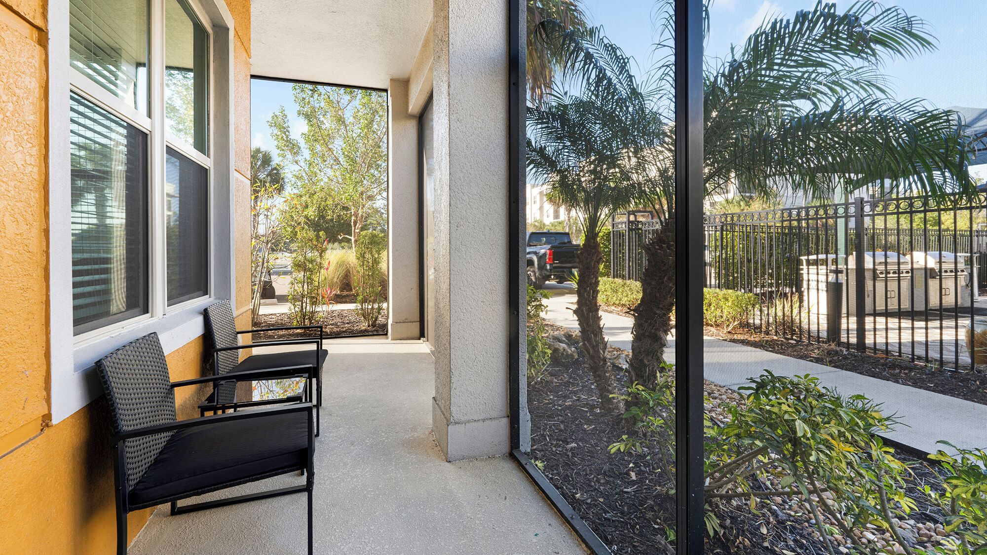 A balcony with a chair, a window, and a view of a street with palm trees and a fence.