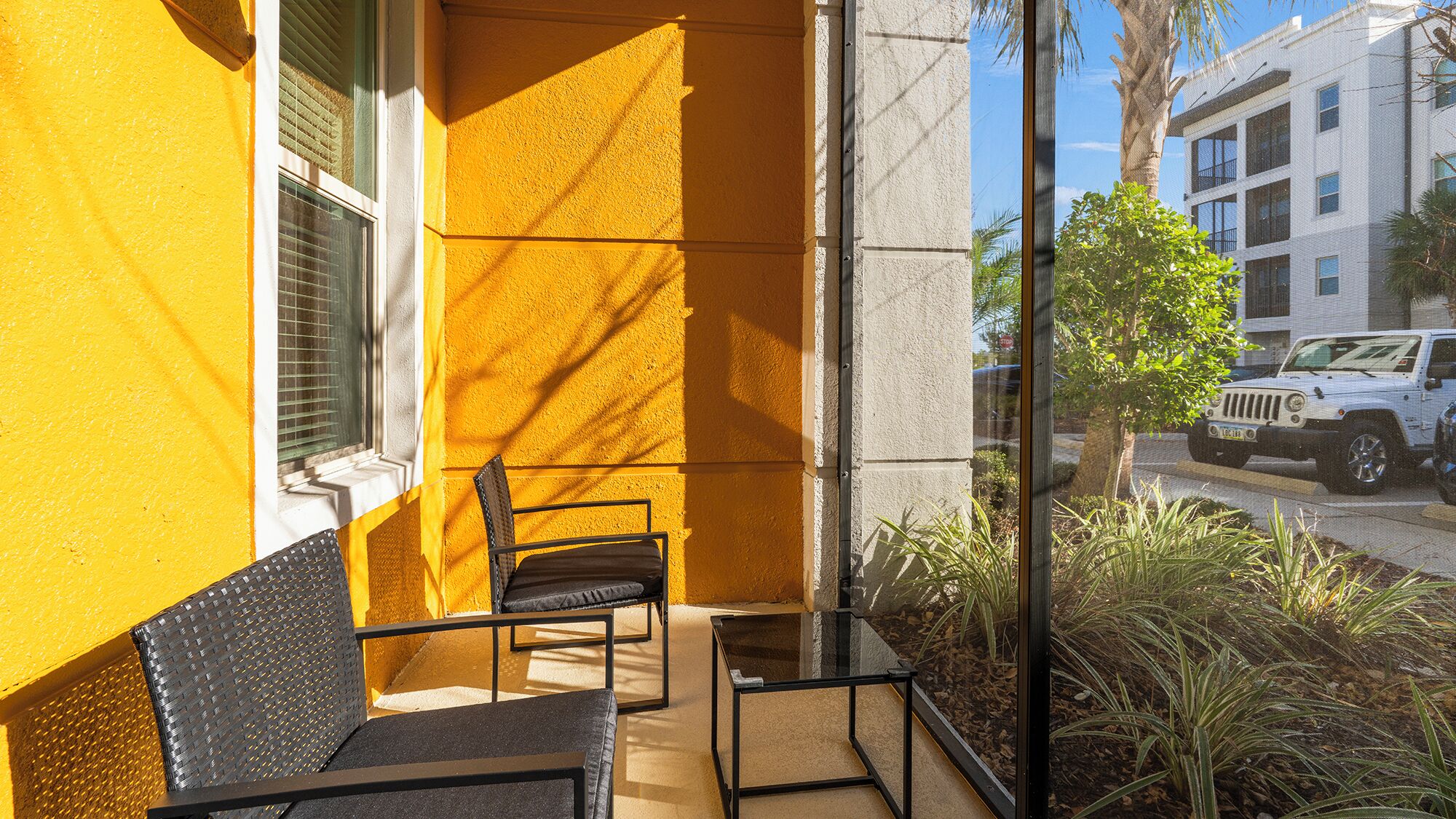 A balcony with two chairs and a table, a yellow wall, and a view of a street with a white vehicle.