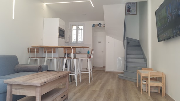 A modern, well-lit living area with a wooden dining table, chairs, a sofa, and a staircase.