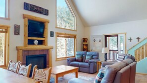A cozy living room with a fireplace, a sofa, a dining table, and a staircase. - Master ensuite  | fenced-in yard | firepit & grill | Lazy Dog Cabin (Angel Fire)