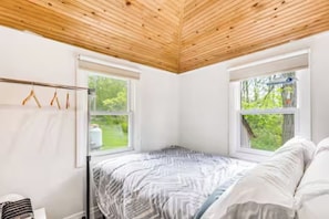 Un dormitorio con techo de madera, dos ventanas, una cama con ropa de cama estampada y un perchero.