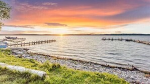 Vista al agua con un muelle que se adentra en el agua, un área verde y un atardecer al fondo.