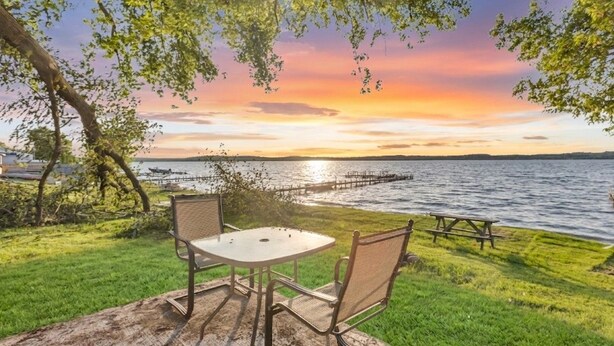 Mesa y sillas en una terraza de madera con vista a un lago al atardecer.