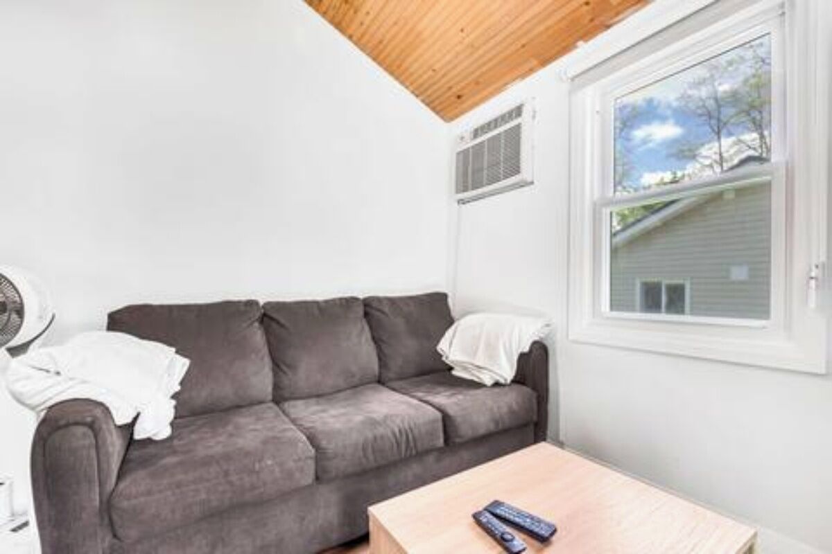 Una habitación con sofá gris, una mesa de centro de madera, una ventana con vistas a una casa y una pared blanca.