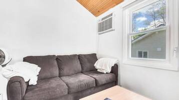 Una habitación con sofá gris, una mesa de centro de madera, una ventana con vistas a una casa y una pared blanca.