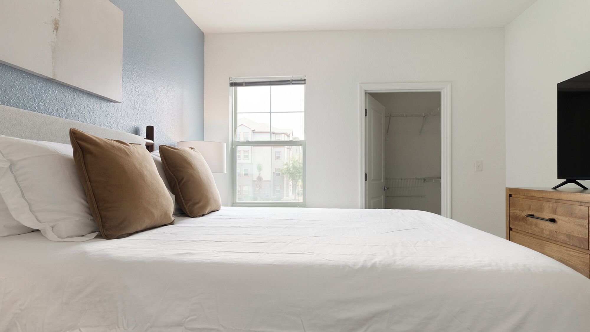 Une chambre à coucher avec un lit, des oreillers, un téléviseur posé sur une commode en bois et une fenêtre donnant sur des immeubles.