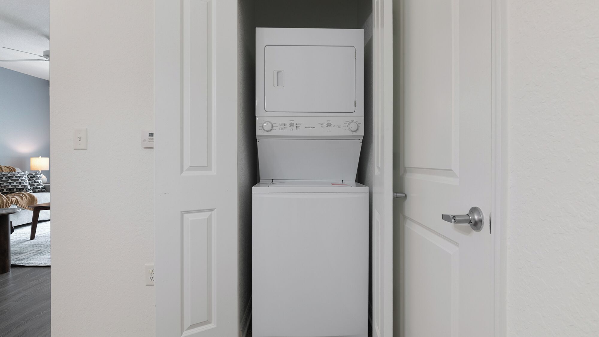 A compact, white laundry room with a built-in washer and dryer, a door with a silver handle, and a visible electrical outlet on the wall.